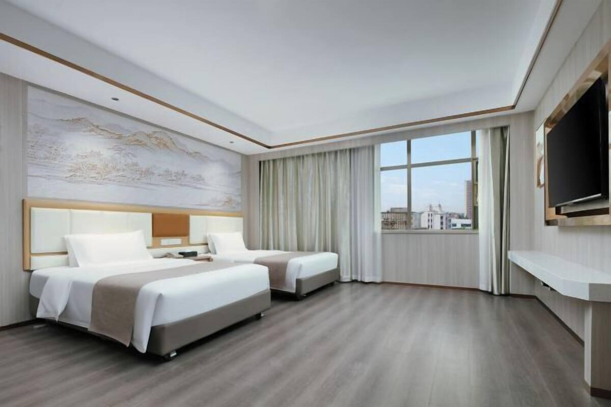 Habitación de hotel con dos camas, un ventanal con cortinas, televisor montado y vista a la ciudad.