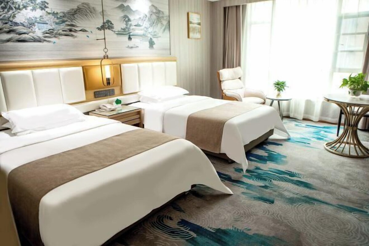A hotel room with two beds, a desk, a chair, a small table with a plant, and a large window.