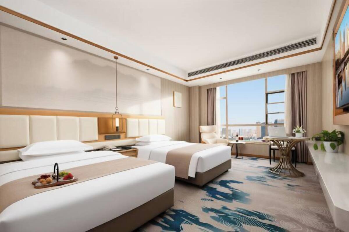 Habitación de hotel con dos camas, zona de comedor, vista a la ciudad y un ventanal grande.