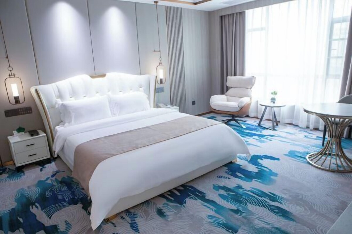 A hotel room with a large bed, a sitting area with a chair and a small table, and a view of the outdoors through the window.