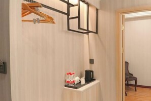 Quarto de hotel com guarda-roupa de madeira, uma pequena mesa com chaleira e garrafas, e uma cadeira no canto.