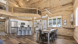 Une cabane en bois spacieuse comprenant un coin repas central, une cuisine et une chambre à coucher située en mezzanine.