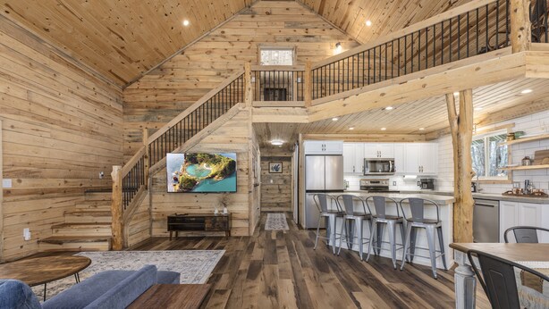 Un vaste espace de vie avec un intérieur en bois, un escalier menant à une mezzanine, une cuisine équipée d’appareils modernes et un téléviseur à écran plat fixé au mur.