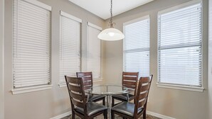 Un coin repas avec une table ronde en verre entourée de quatre chaises en bois, des stores blancs aux fenêtres et un luminaire suspendu.