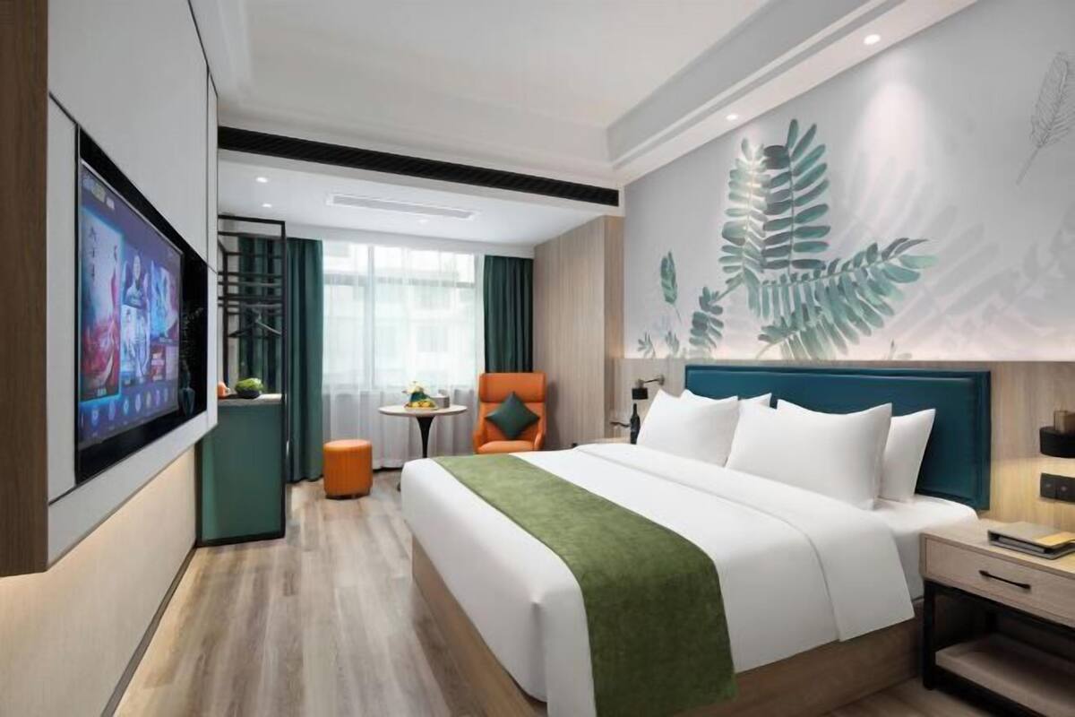Een moderne hotelkamer met een groot bed, een aan de muur bevestigde televisie, een klein tafeltje met een stoel en een decoratieve plant aan de wand.