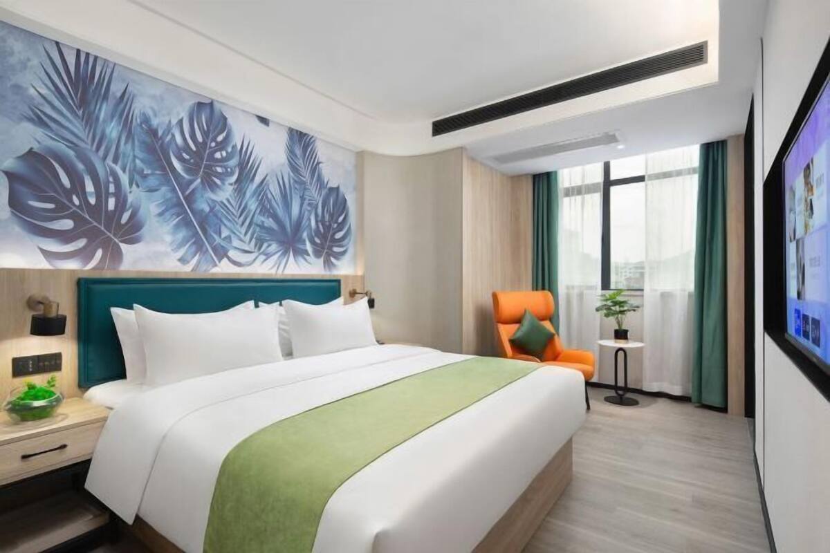 Hotelkamer met groot bed, hoofdeinde, een aan de muur bevestigde televisie, nachtkastje met plant en raam met gordijnen.