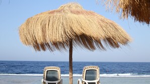 Dos sillas de playa bajo un toldo de paja en una playa arenosa, con el océano al fondo.