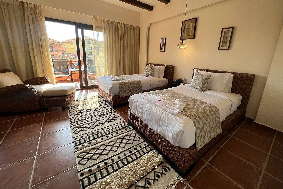 A hotel room with two beds, a sofa, a patterned rug, and a view of buildings outside.