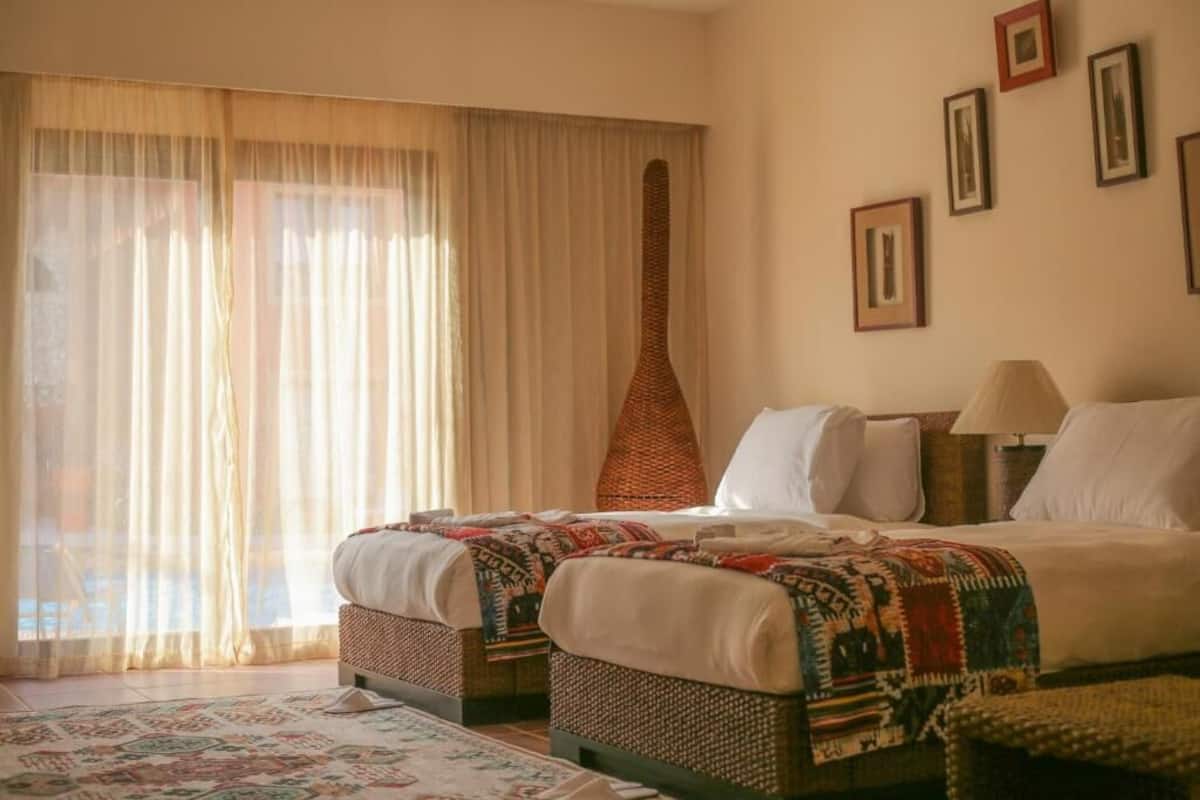 A hotel room with two beds, a large window with sheer curtains, a woven chair, and a patterned rug.