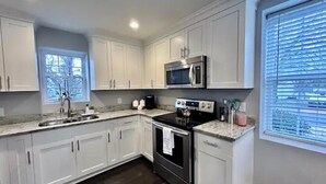 A modern kitchen with white cabinets, a stainless steel microwave, a black stove, and a window with blinds. - Relaxing Stay on Trade Street (Greer)
