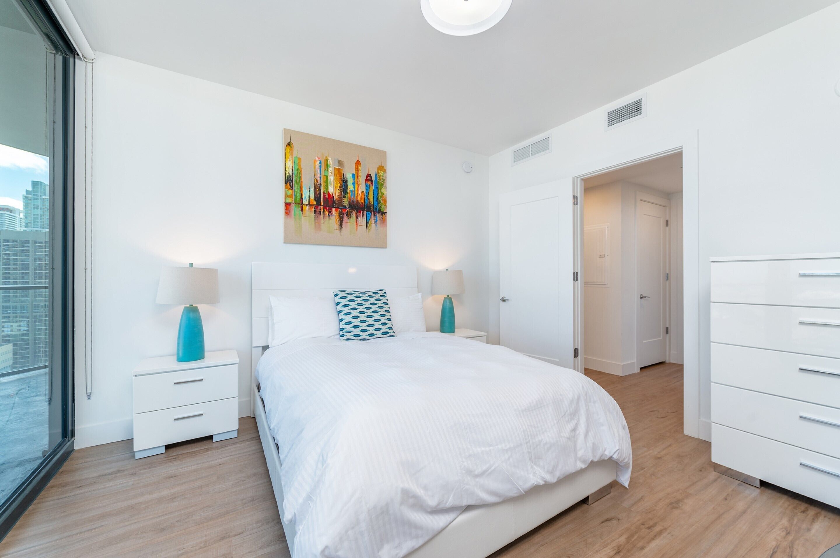 A modern bedroom with a large bed, white walls, and a cityscape painting.