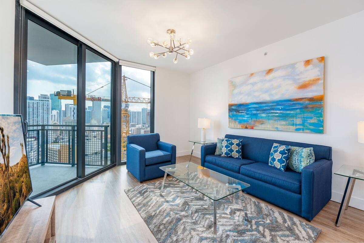 A modern living room with a blue sofa, a glass coffee table, and a view of a cityscape through large windows.