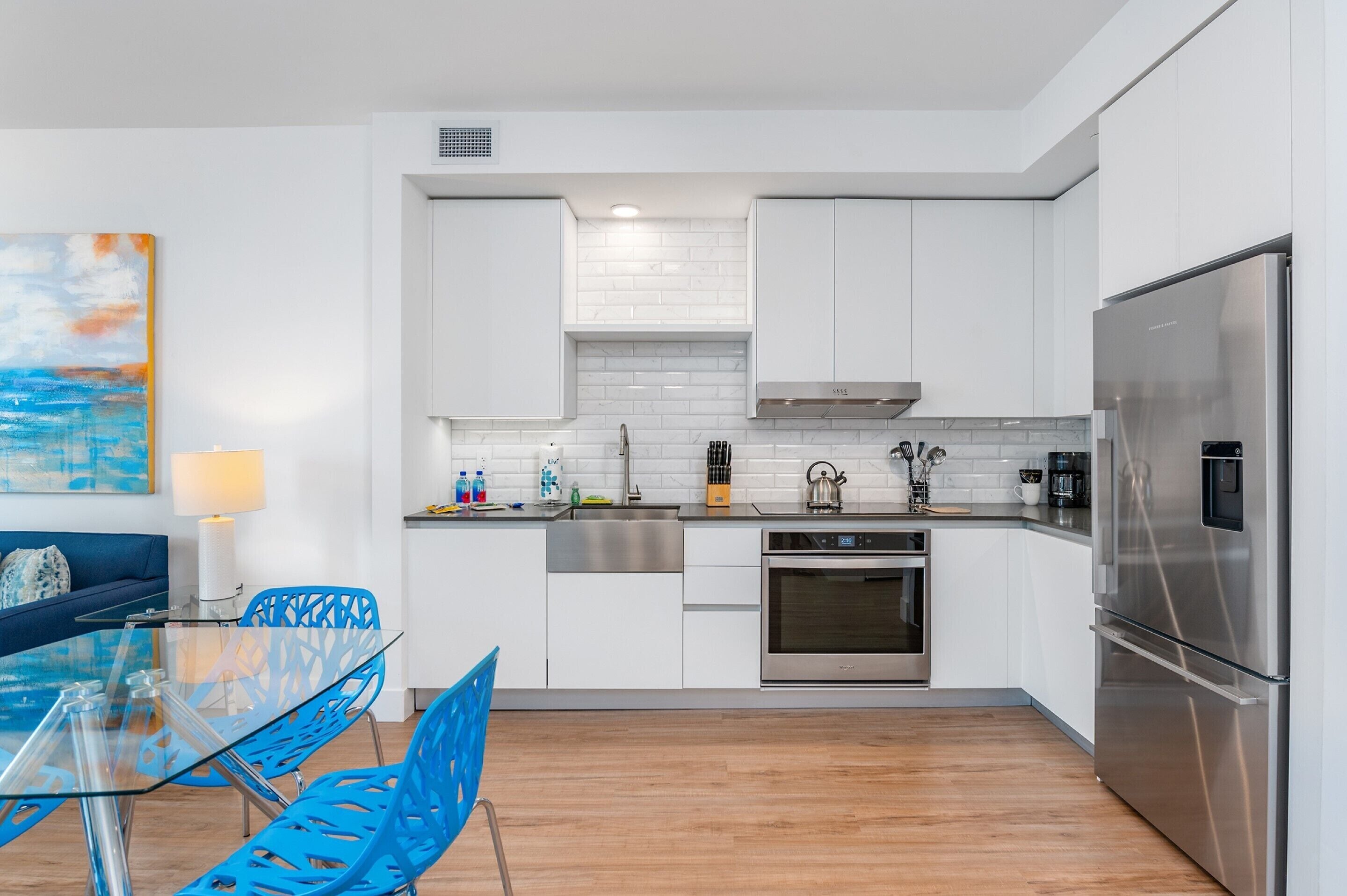 A modern kitchen with white cabinets, stainless steel appliances, and a glass dining table with blue chairs.