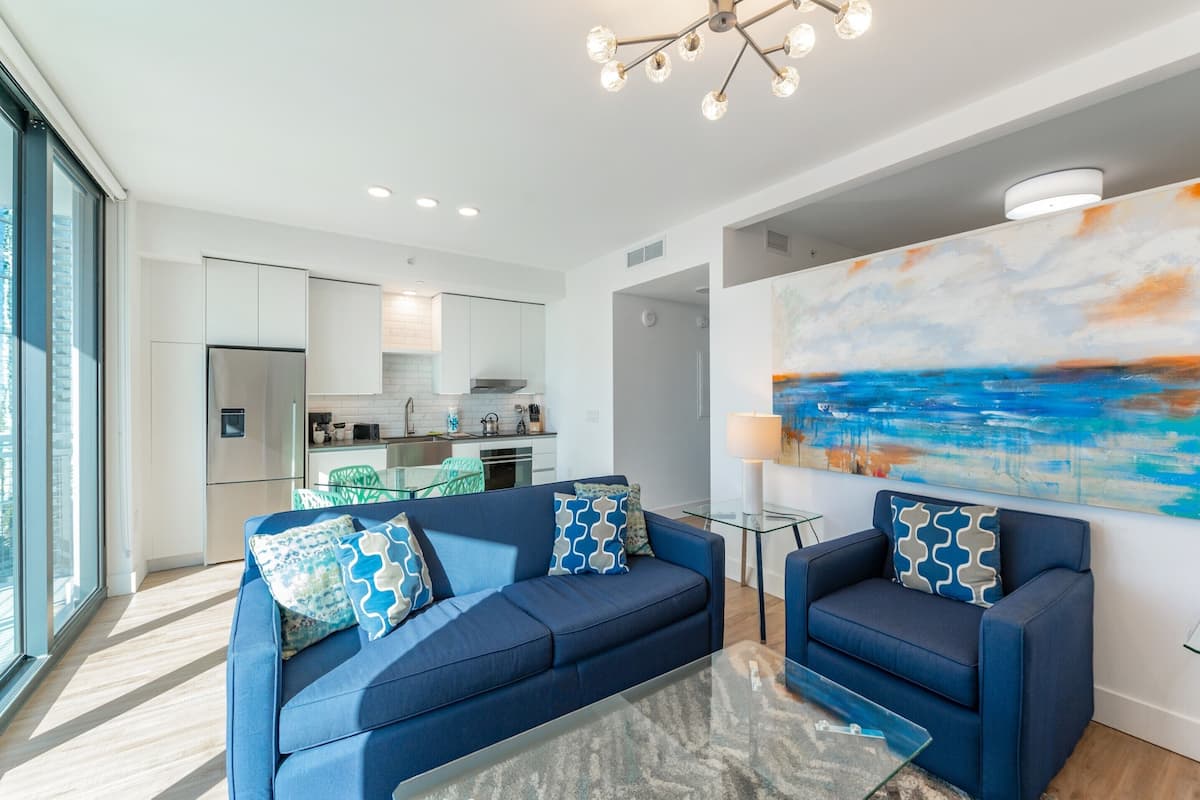A modern living room with a blue sofa set, a glass coffee table, and a large abstract painting on the wall.