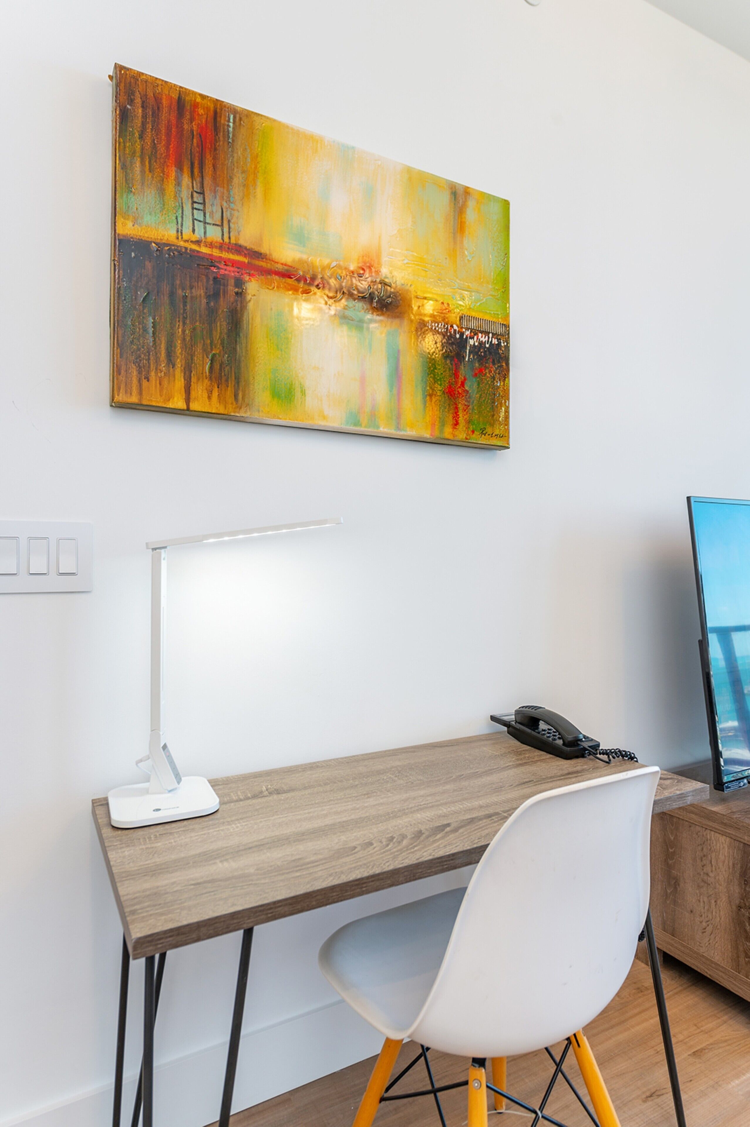 Ein moderner Arbeitsplatz mit Schreibtisch aus Holz, einem weißen Stuhl, einer Schreibtischlampe und einem Bild an der Wand.