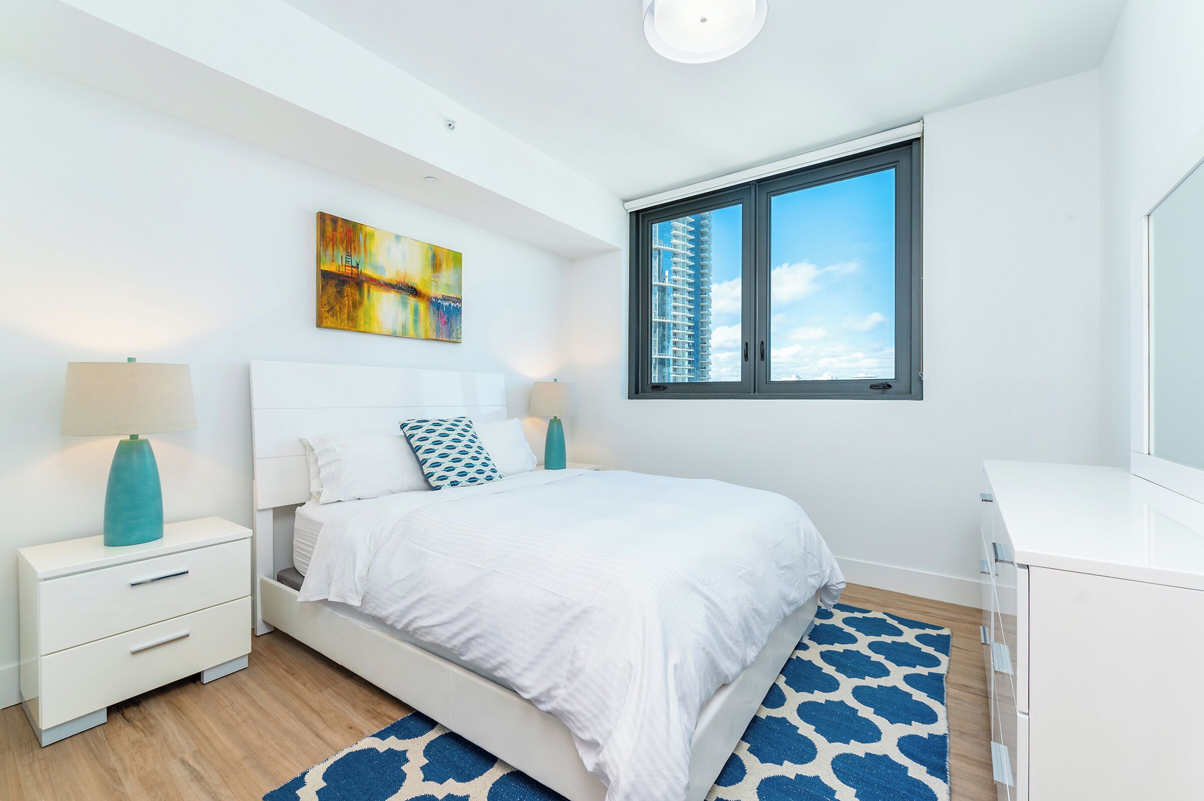 Een moderne slaapkamer met een bed, nachtkastjes, een dressoir, een raam met uitzicht op een gebouw en een schilderij aan de muur.
