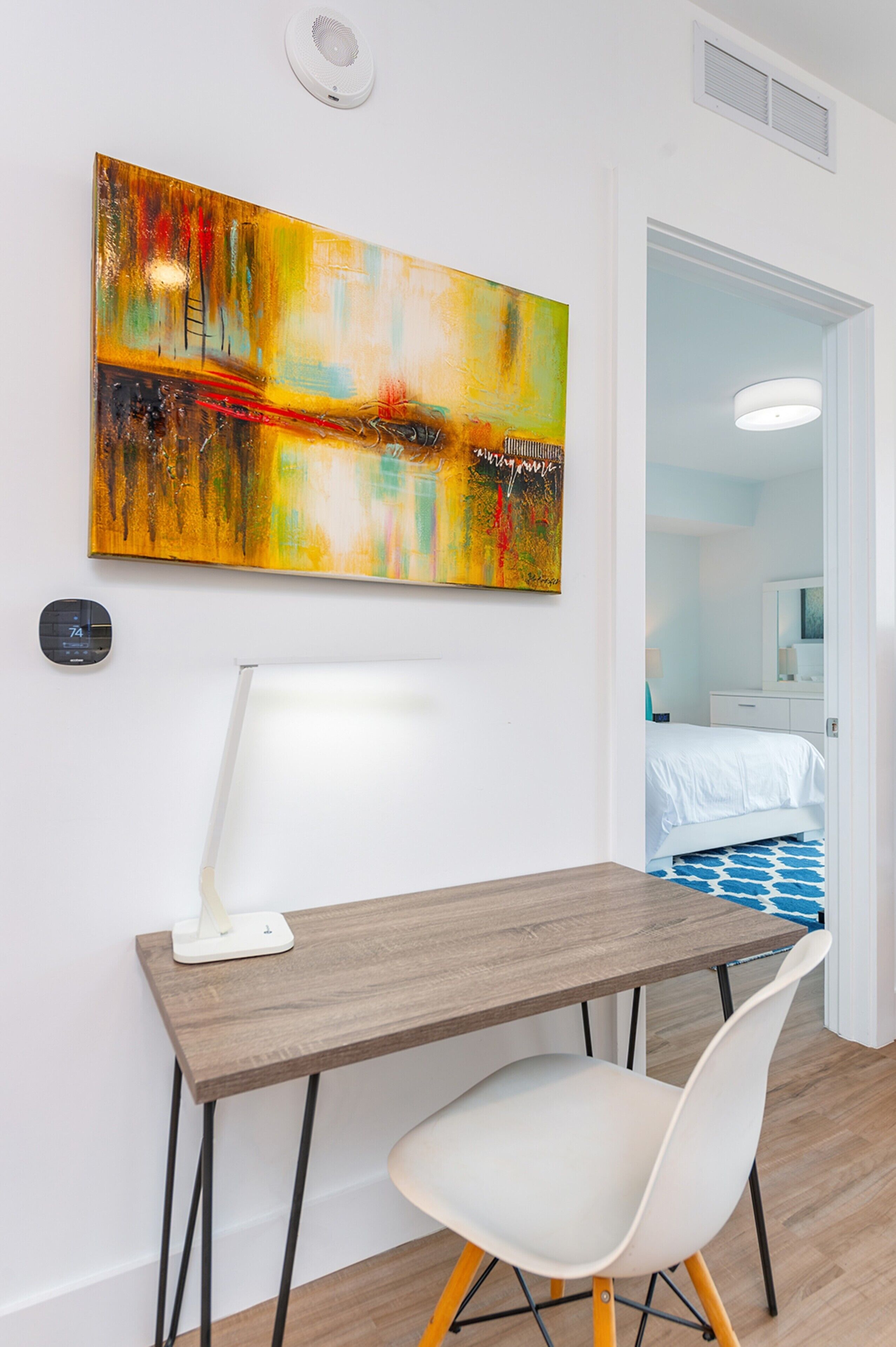 Una habitación moderna con un escritorio de madera, una silla blanca y un cuadro abstracto colorido en la pared.