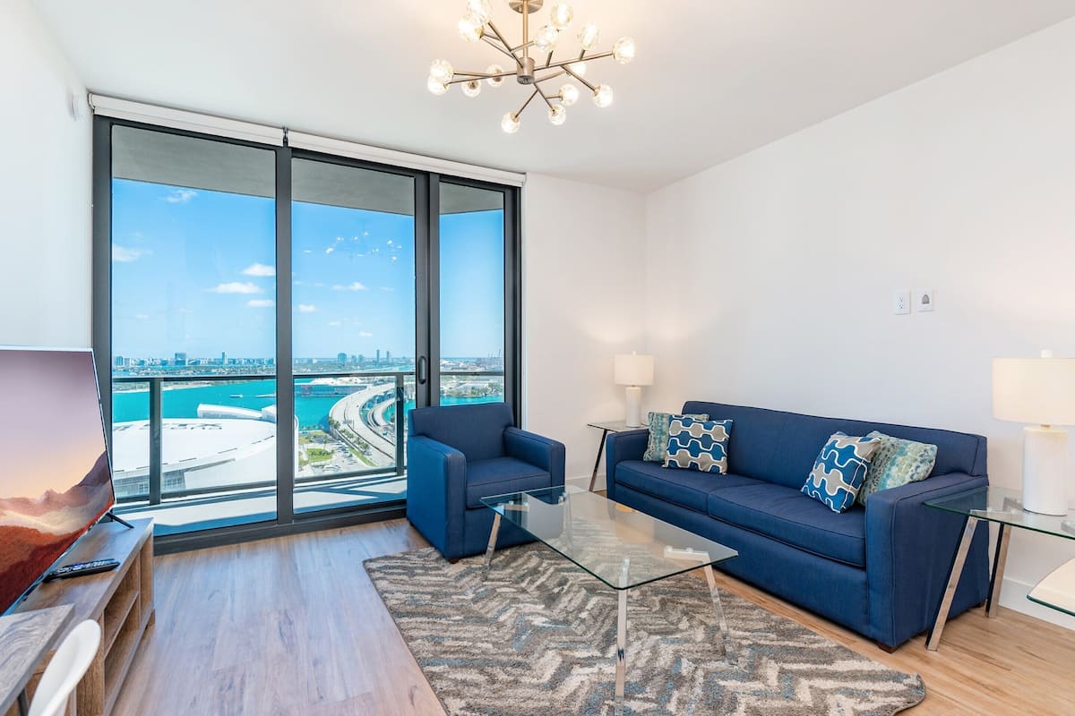 A modern living room with a large window, a blue sofa, a glass coffee table, and a view of a cityscape and water.