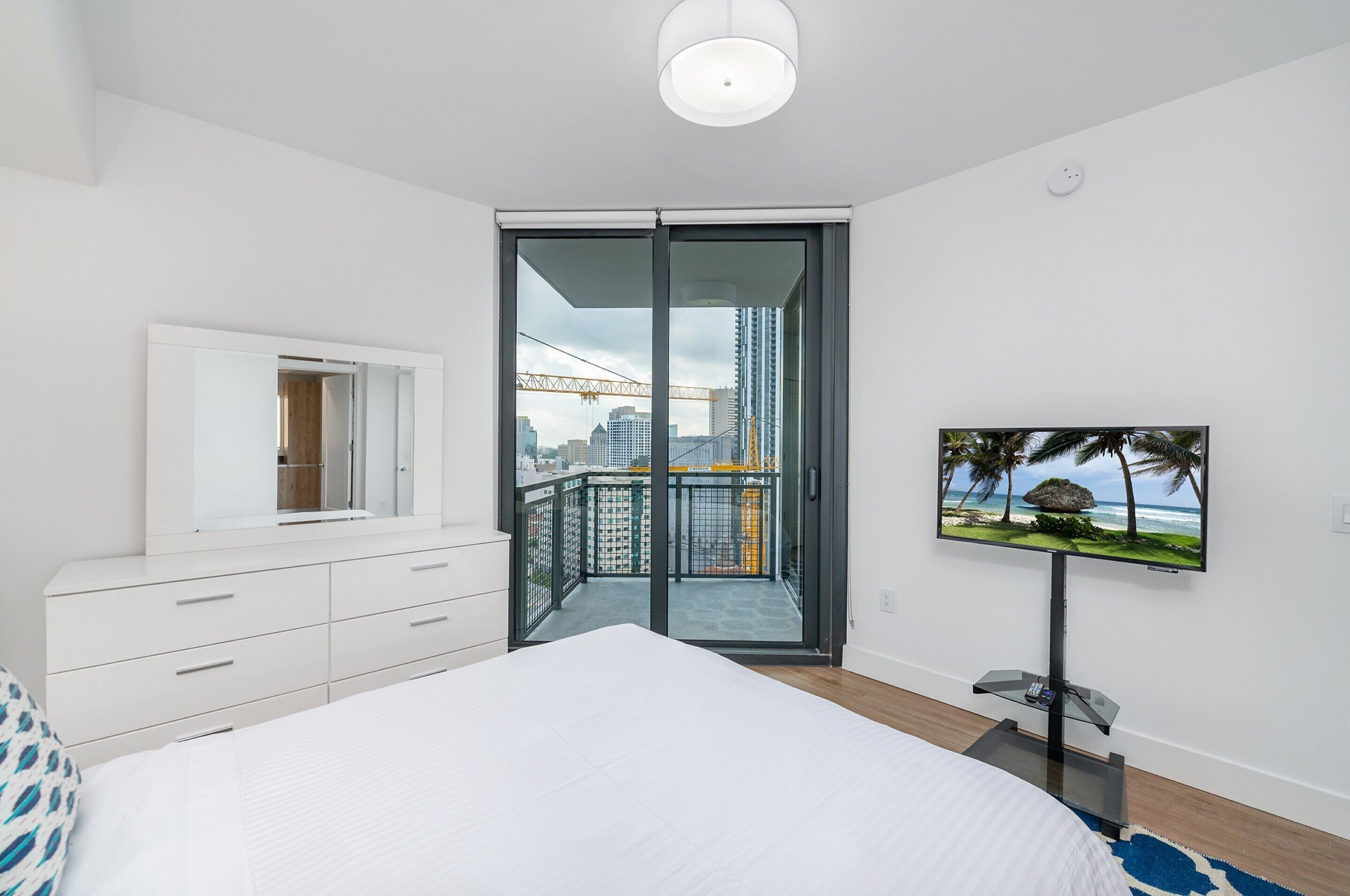 A bedroom with a large bed, a dresser, a TV on a stand, and a balcony with a city view.