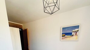 Un dormitorio con una cama, una manta rosa, un cuadro enmarcado con una escena de playa y una lámpara con diseño geométrico.