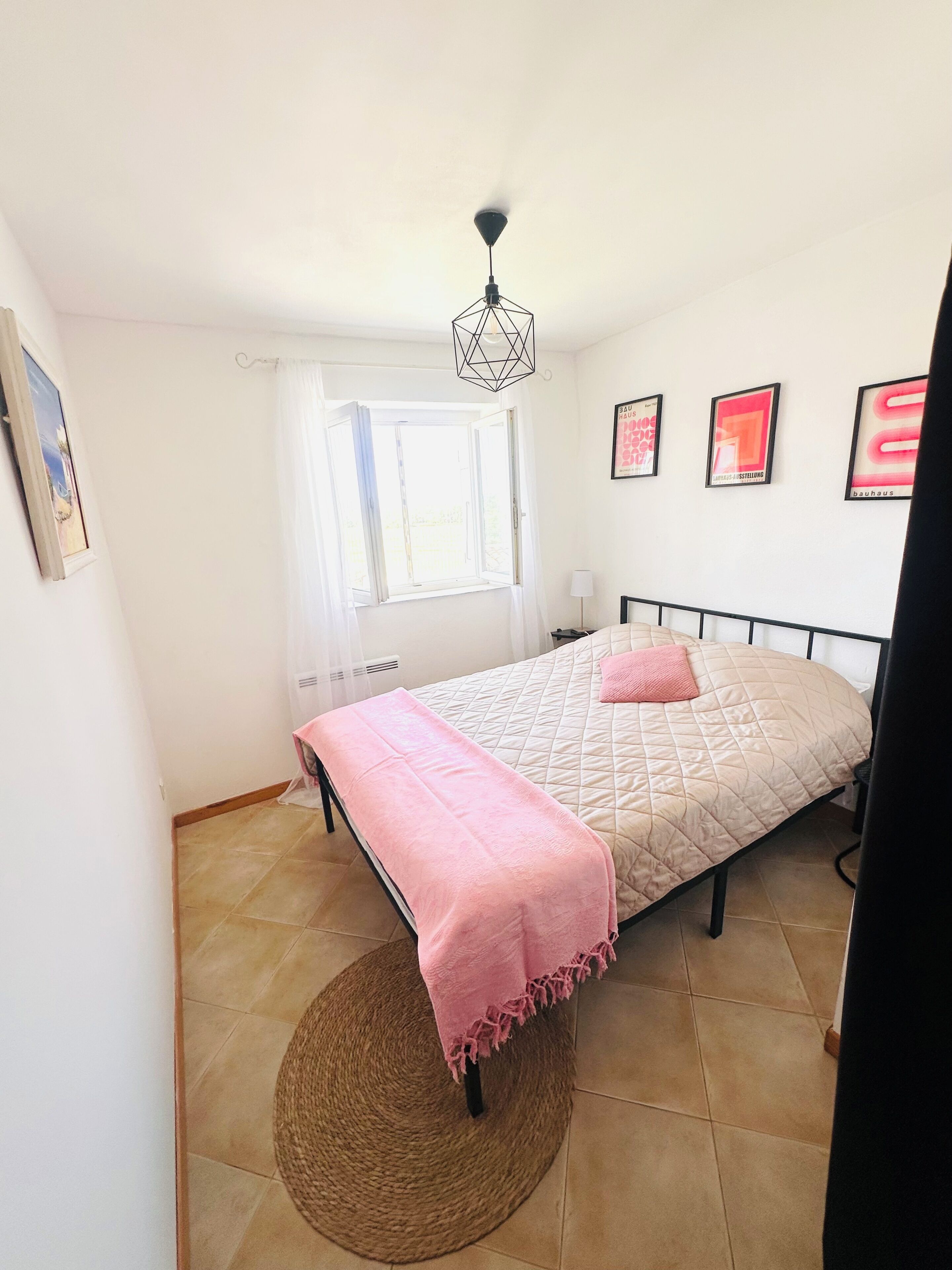 Ein Schlafzimmer mit einem Bett, einem Fenster, einer Pendelleuchte und gerahmten Bildern an der Wand.