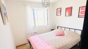 Ein Schlafzimmer mit einem Bett, einem Fenster, einer Pendelleuchte und gerahmten Bildern an der Wand.