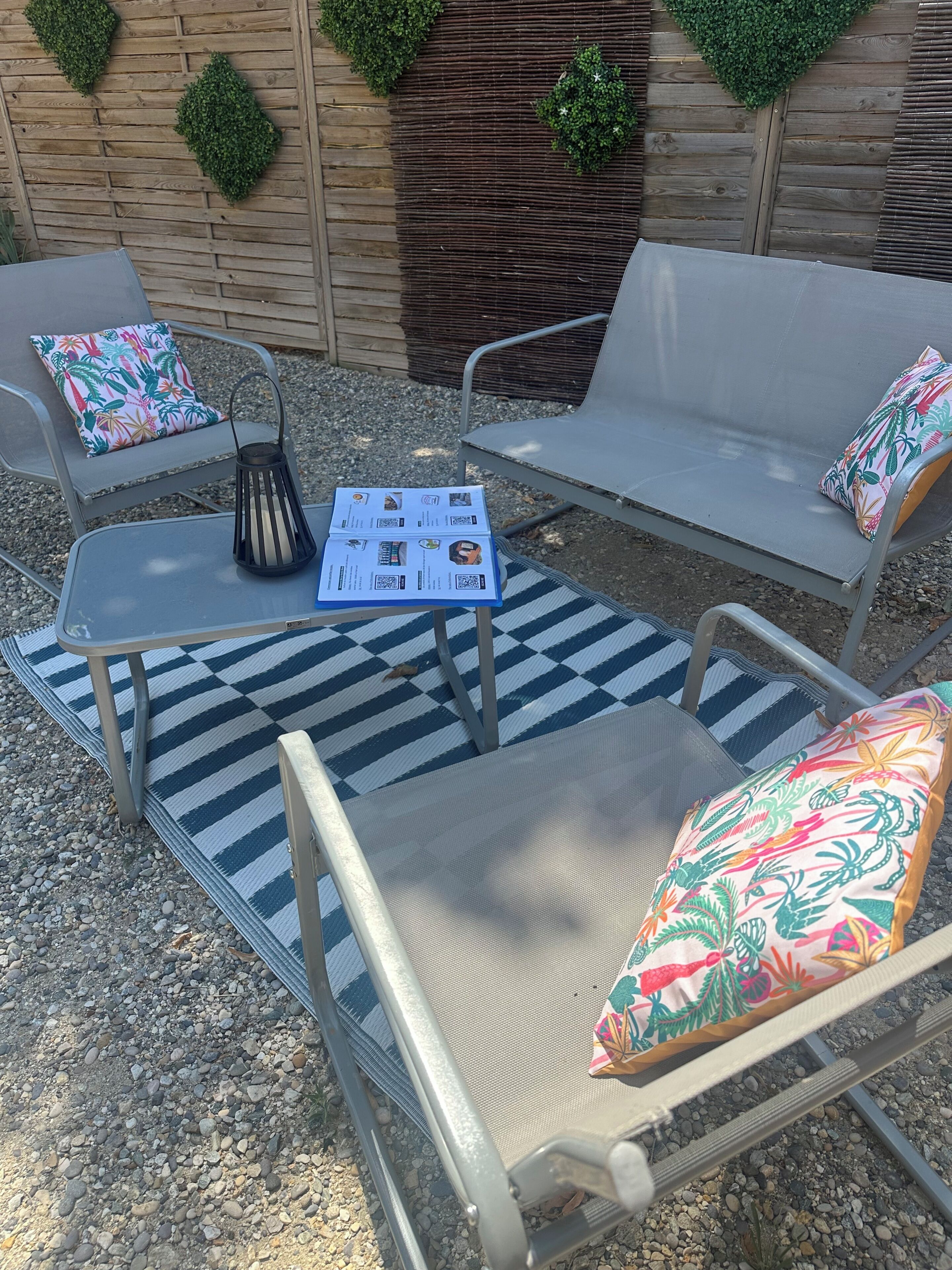 Een buitenzitgedeelte van metaal, met een geruit kleed, een tafel met een QR-code en twee stoelen met gestreepte kussens.