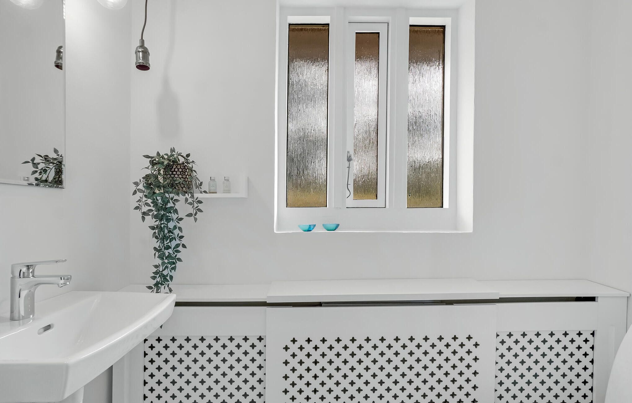 Une salle de bain avec un lavabo blanc, un miroir, une fenêtre à vitre dépolie et une plante.