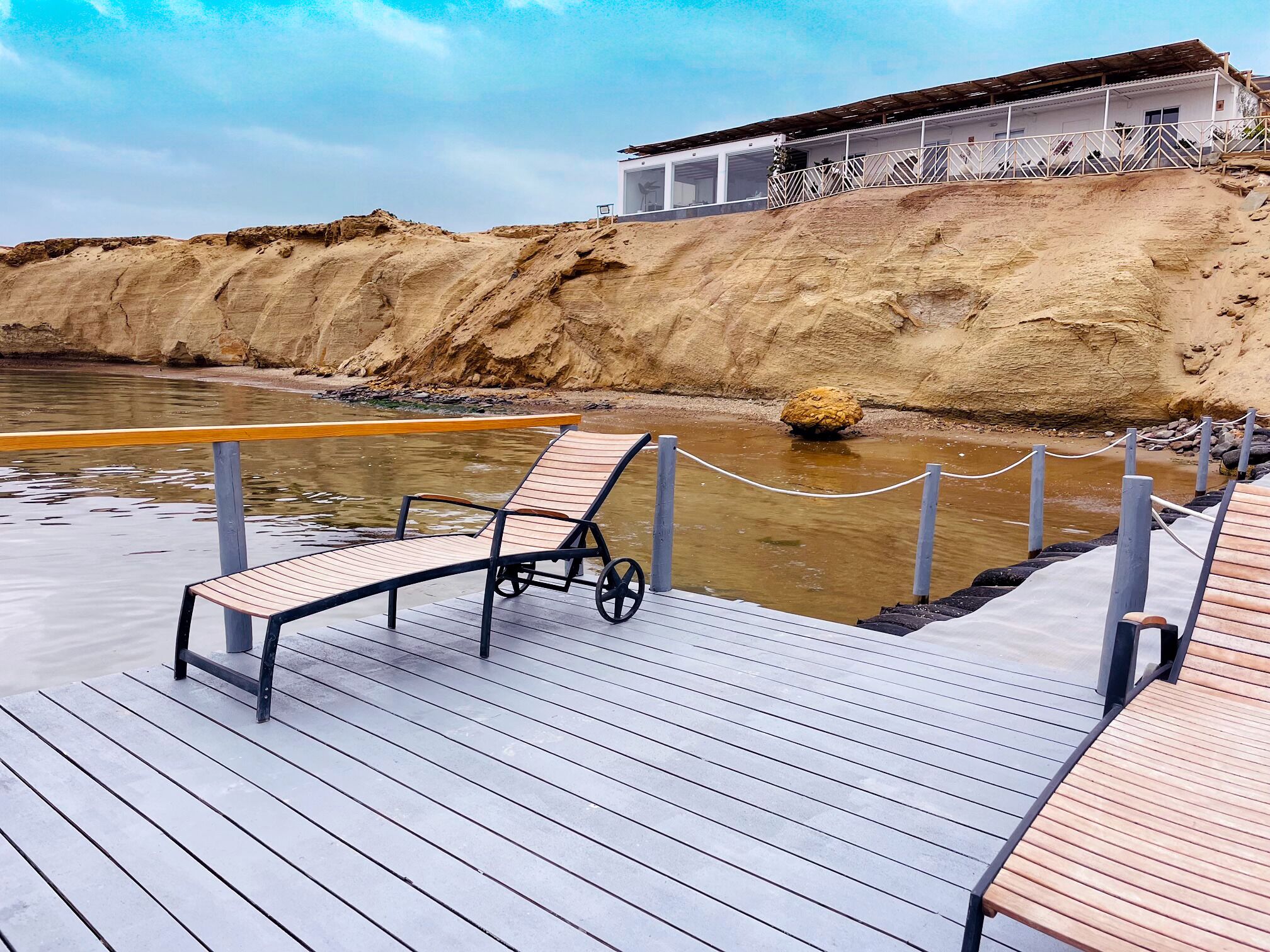 A wooden deck with lounge chairs and a wheelchair, overlooking a river with a sandy bank and a building in the background.
