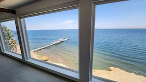 A room with large windows offering a view of a calm sea, a wooden deck, and a sandy beach. - HOTEL BOUTIQUE ATENAS  (Paracas)