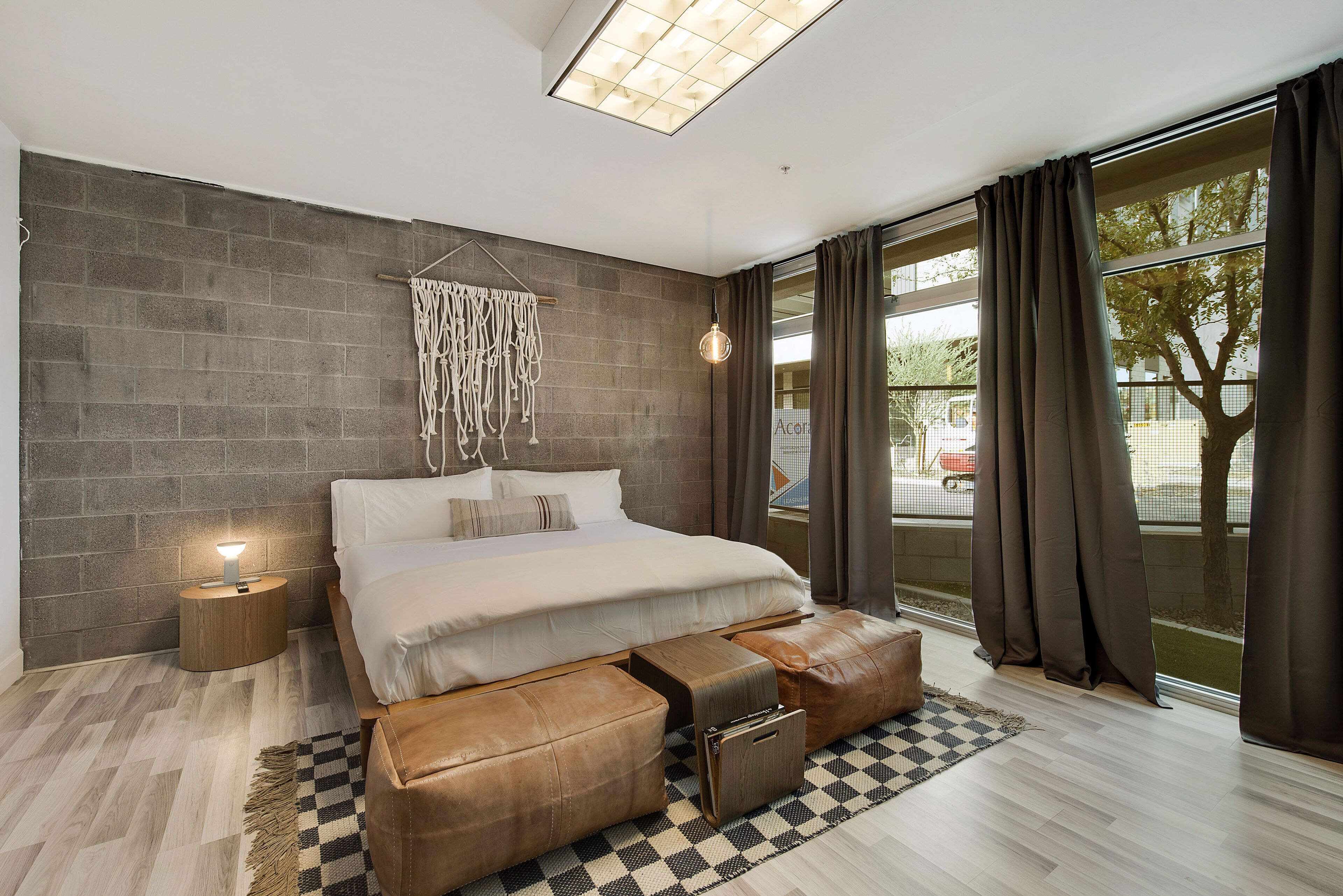 Une chambre moderne avec un grand lit, une table de chevet avec une lampe, une décoration murale en macramé et une vue sur l’extérieur grâce à de grandes fenêtres.