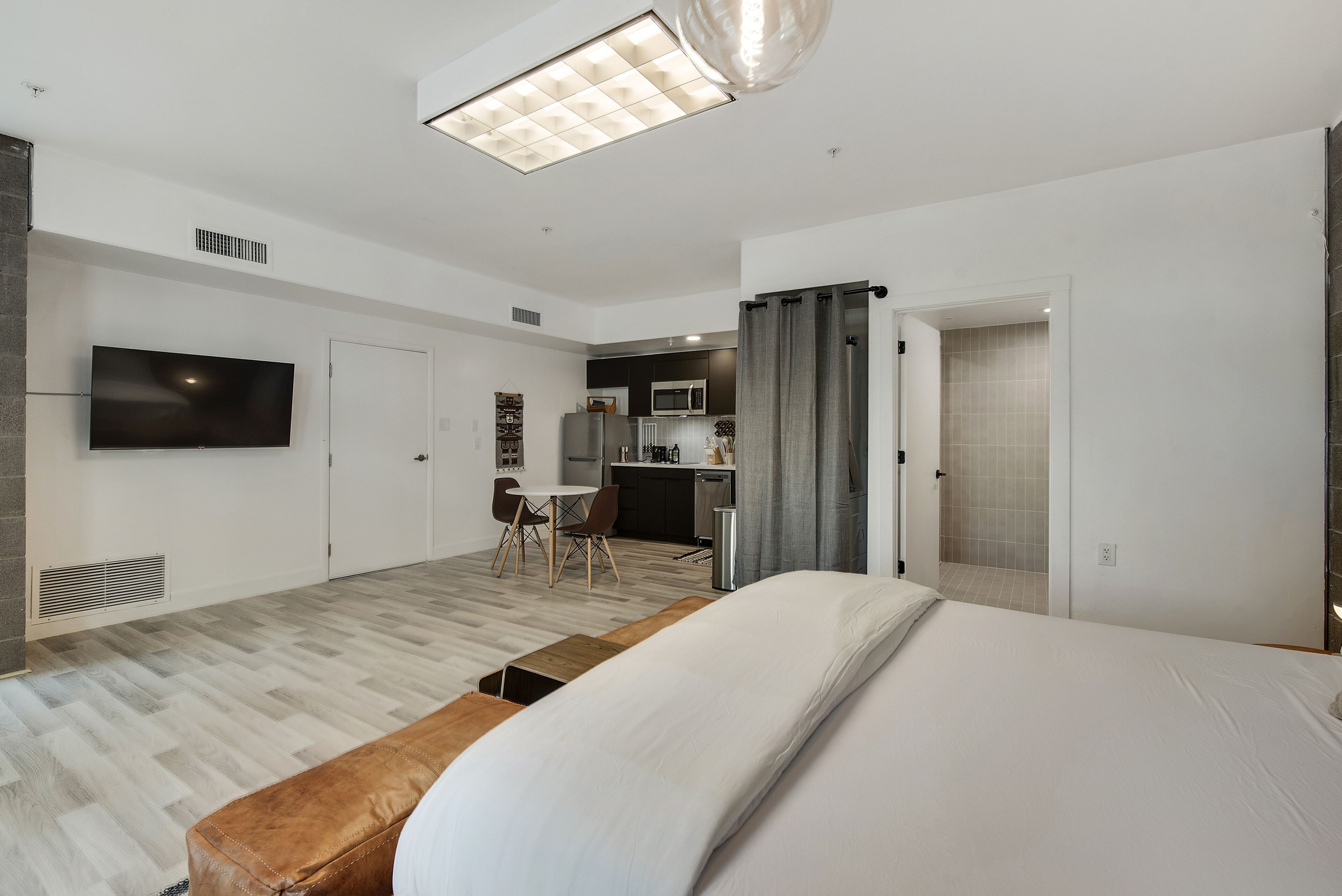 A modern hotel room with a large bed, a TV mounted on the wall, a small kitchenette, and a dining area with chairs.