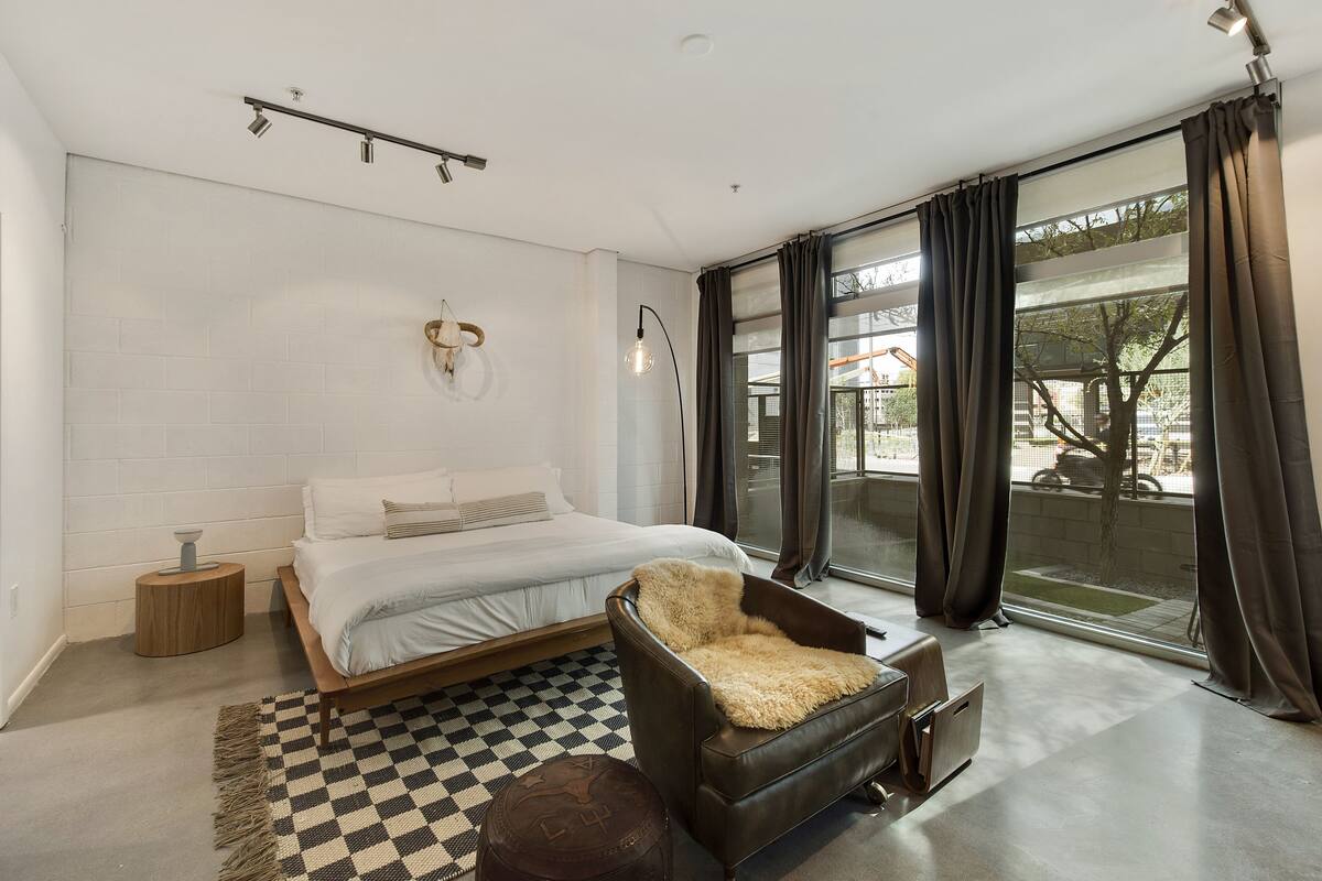 Un dormitorio moderno con una cama grande, un sillón de cuero, un tapete a cuadros y vistas al exterior a través de amplios ventanales.