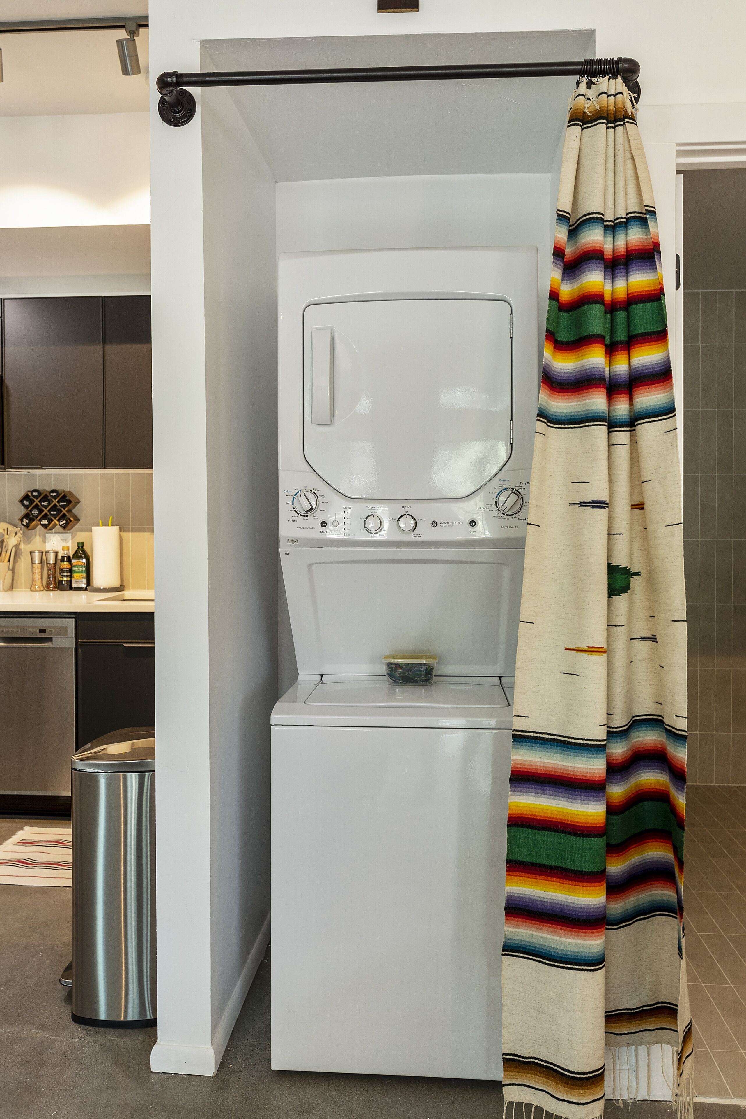 A compact laundry room with a washing machine and dryer, a metal trash can, and a striped shower curtain.