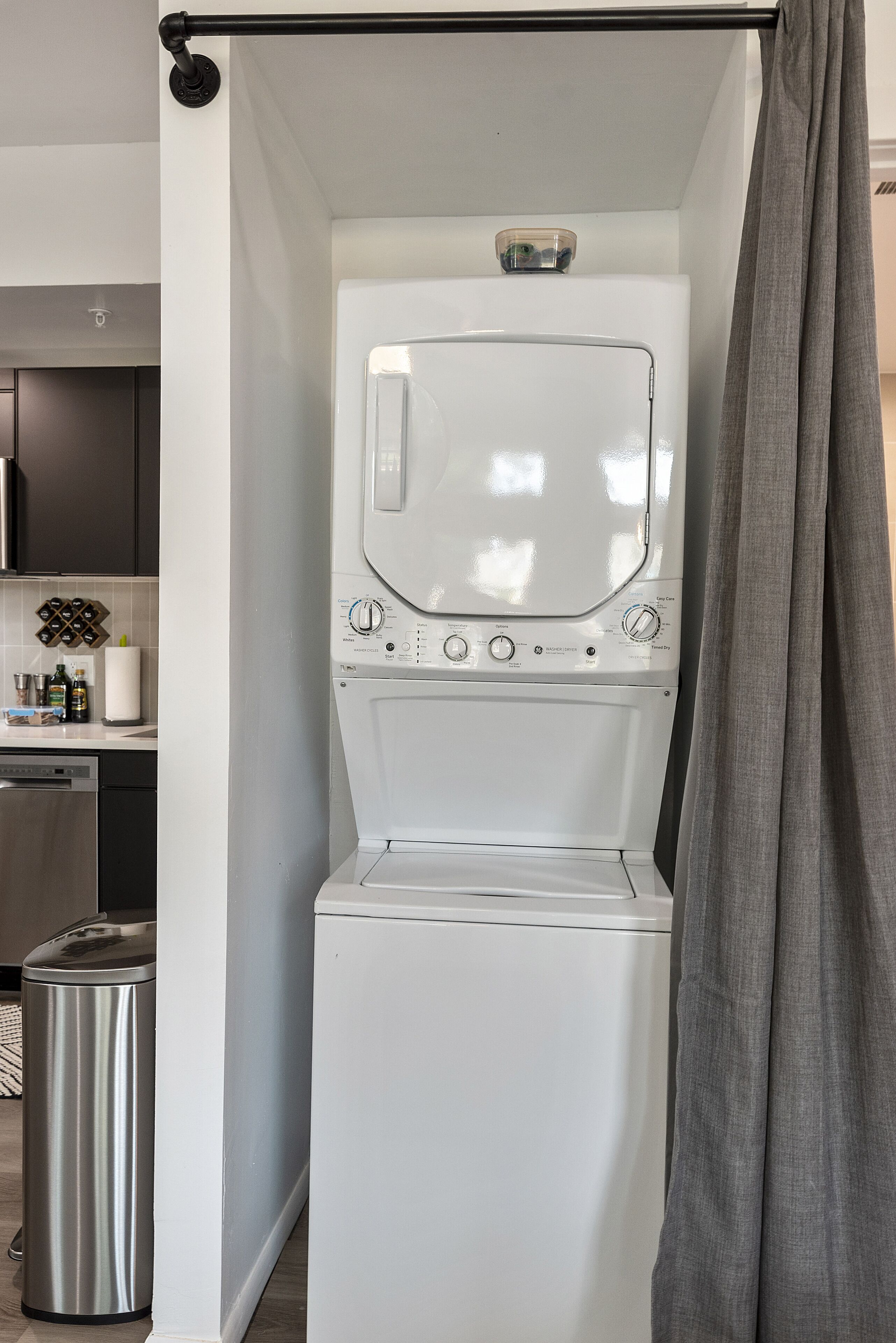 Une petite buanderie moderne, dotée d’un lave-linge et d’un sèche-linge encastrés, d’une poubelle en acier inoxydable et d’un coin cuisine visible au fond.