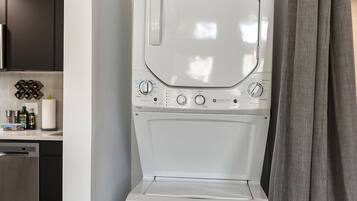 Une petite buanderie moderne, dotée d’un lave-linge et d’un sèche-linge encastrés, d’une poubelle en acier inoxydable et d’un coin cuisine visible au fond.