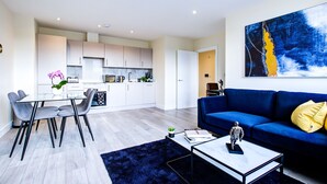 A modern living room with a blue sofa, a glass dining table with chairs, a coffee table, and a kitchen in the background. - Charming 2-bed Apartment in Cheam, Sutton (Sutton)