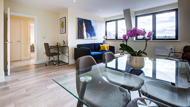 A modern living room with a glass dining table, comfortable seating, a sofa with a yellow pillow, and a large window with a view. - Charming 2-bed Apartment in Cheam, Sutton (Sutton)
