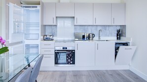 A modern kitchen with white cabinets, a built-in oven, a microwave, and a glass dining table. - Charming 2-bed Apartment in Cheam, Sutton (Sutton)