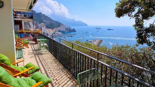 Ein Balkon mit grünen Sitzmöbeln, gefliestem Boden und Blick auf das Meer mit Booten und einer Burg am Hügel.