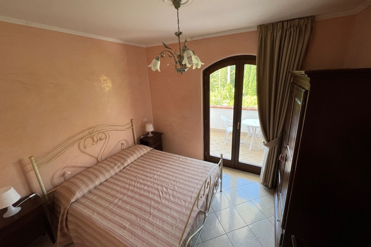 Ein Schlafzimmer mit Bett, Nachttisch, Lampe, Deckenventilator und Blick ins Freie durch eine Tür.