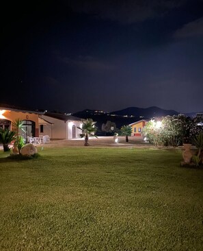 A well-lit outdoor area with a grassy lawn, a patio with outdoor furniture, and a building with a visible entrance. - Villa Lia Cilento (Giungano)