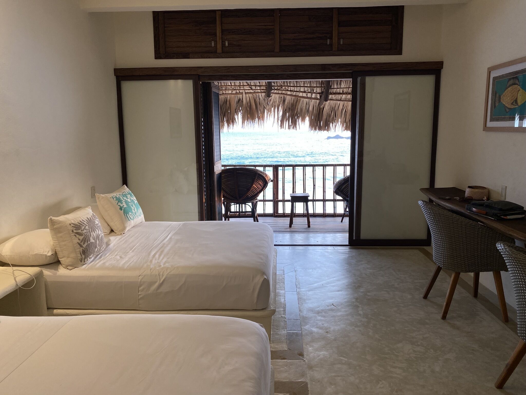 A hotel room with two beds, a desk, and a balcony with a view of the sea.