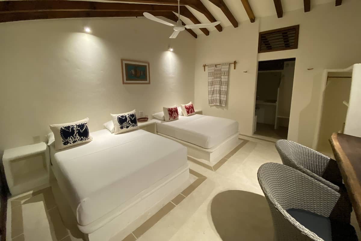 Una habitación con dos camas, un ventilador de techo, un cuadro y una puerta que da al baño.