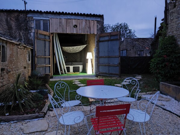 Un área de estar al aire libre de estilo rústico con una mesa y sillas, rodeada por un muro de piedra y vegetación.