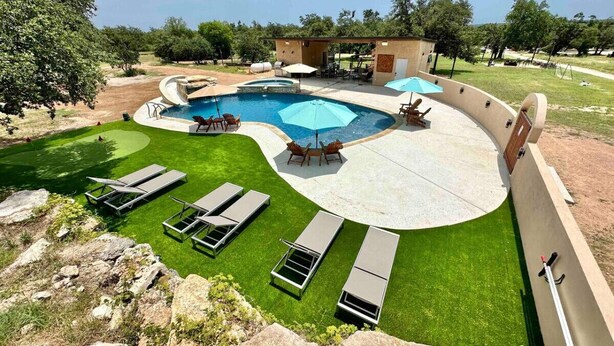 A swimming pool with a curved edge, surrounded by a grassy area with lounge chairs and a small building with an umbrella. - Tree Top Retreat-Pool-Playground-Brewery (Dripping Springs)
