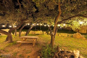 Un espace pique-nique avec un banc en bois et une table à pique-nique sous un arbre, entouré de rochers et illuminé par des guirlandes lumineuses.