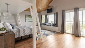 A bedroom with a loft bed, wooden ceiling beams, a large window with curtains, a painting on the wall, and a small table with a plant.