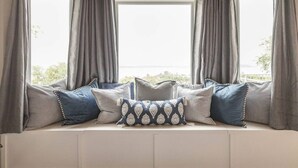 A bay window with grey curtains, a white cushioned bench, and several pillows.