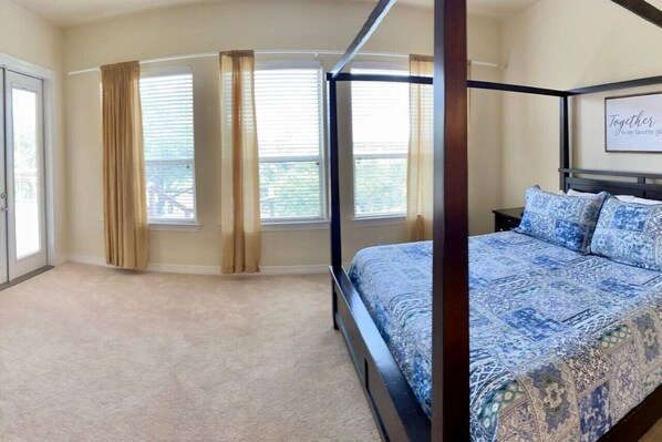 Un dormitorio con cama con dosel, alfombra color beige, dos ventanas con cortinas y una cita enmarcada en la pared.